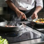 Picture of two frying pans on a commercial kitchen cooker, with the hands of a male chef attending to one of them.
