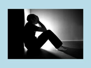 Silhouette of a lonely person sat against a wall, with their head in their hands.