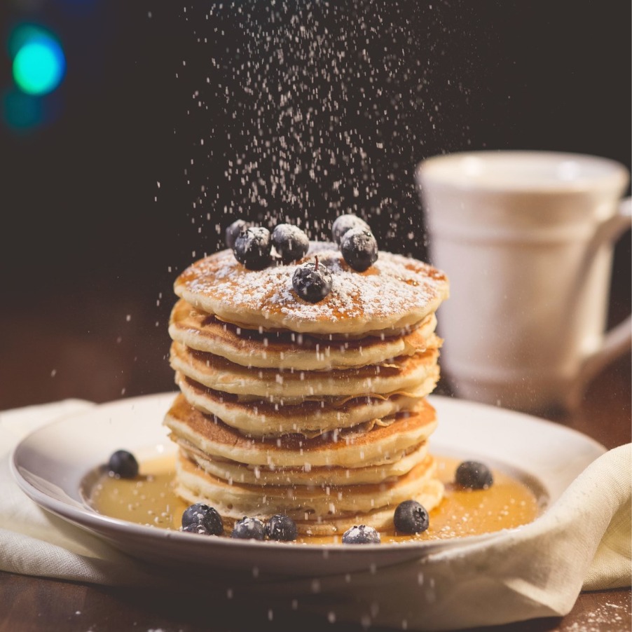 A stack of American-style pancakes with blueberries on top, and icing sugar being dusted on top.